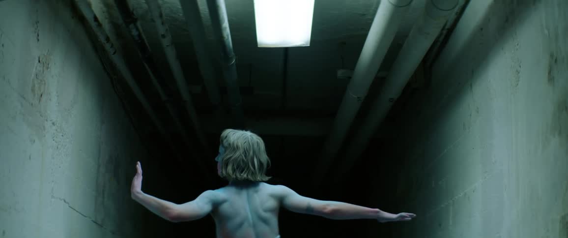 A young woman covered in blue powder takes dramatic poses in a concrete corridor