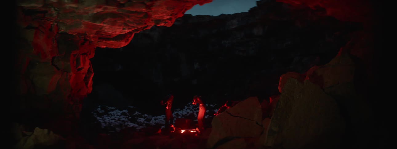 Two women and a man gather around a glowing object on the ground of a cave at night