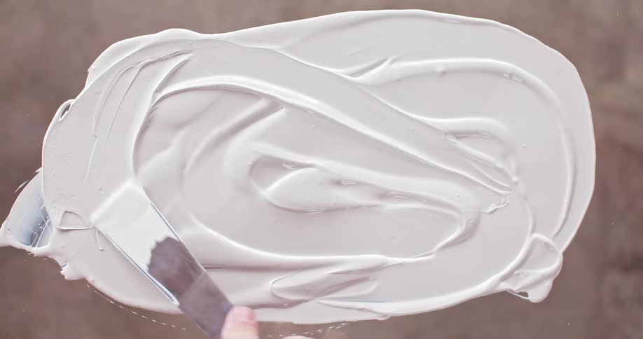 A person spreads white cream with a metallic spatula on a table top