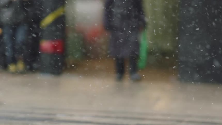 Pedestrians walking on a sidewalk while snow falls and cars drive past