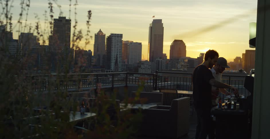 Two men standing on a building patio one cooking on a barbecue pit with buildings and the sky in the background