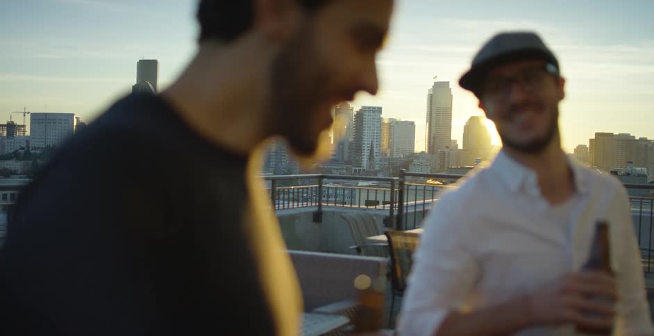 A pair of men drink beer from bottles on the rooftop of a building during the day
