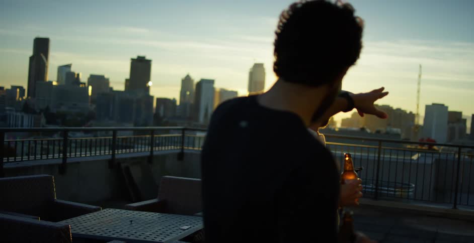 Two male friends drinking beer on the rooftop admiring the skyline at dusk