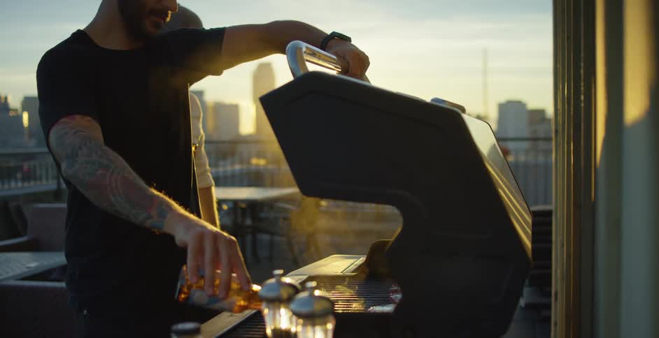 A man grilling on a rooftop in a city pours beer over the food on the grill