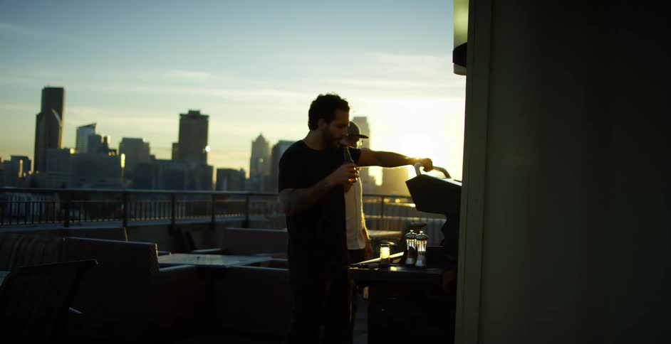 Friends drinking beer while grilling on roof top overlooking skyline at dusk