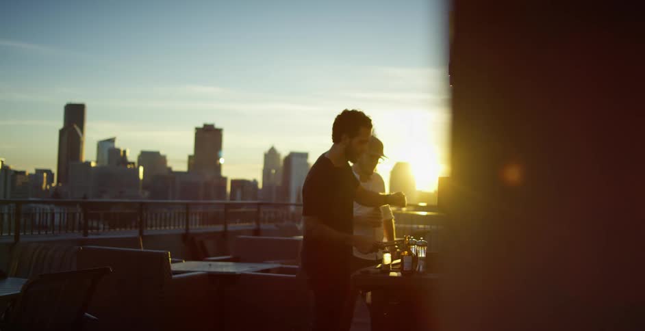 A pair of men drink beer and grill food on the rooftop of a building as the sun shines between the skyscrapers of the city in the background