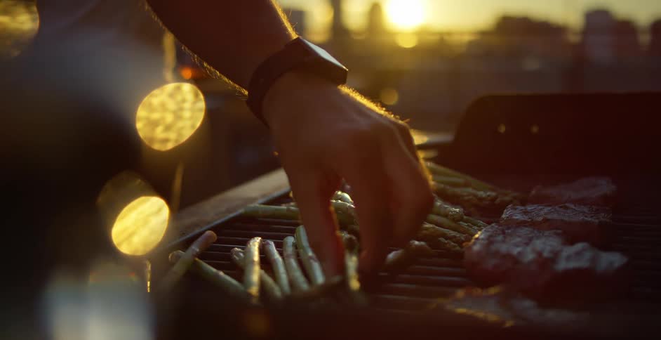 A man cooks on a grill as his friend stands next to him and drinks a beer