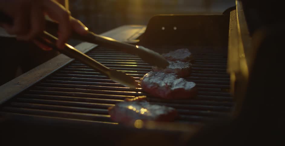 A man uses metal tongs to rearrange steaks on a grill as he cooks on the rooftop of a skyscraper in a metropolis with his friend