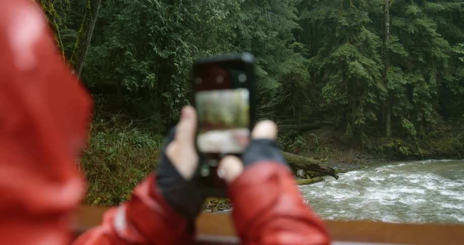 A person in a red raincoat stands on a wooden bridge and uses a smartphone to take photos of the river that runs beneath it
