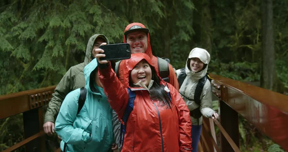 A group of hikers in raincoats pose for a selfie on a wooden bridge in a forest on a rainy day