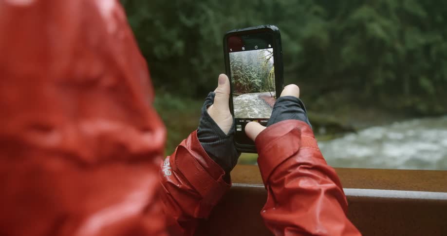 A person with a raincoat and a smart phone taking a picture of a river and trees