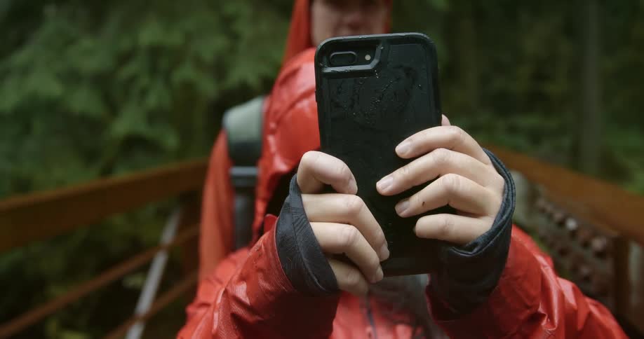 A young asian woman in a red raincoat uses her smartphone to take a selfie with a man in a matching raincoat while standing on a wooden bridge in a woodland on a rainy day