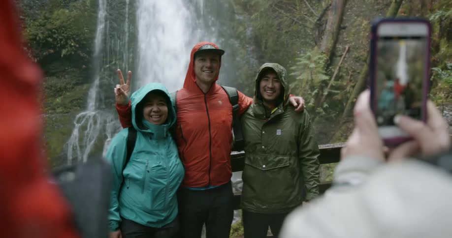 Three hikers pose on a wooden bridge in front of a waterfall as two other hikers use their smartphones to take photos