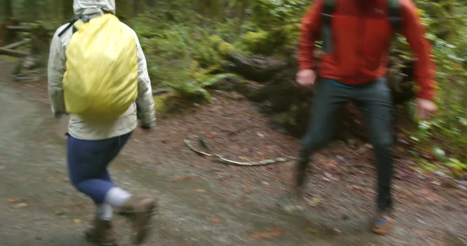 A pair of hikers run through the rain on a dirt path and attempt to splash each other by stomping in the puddles of water as another pair of hikers standby and watches