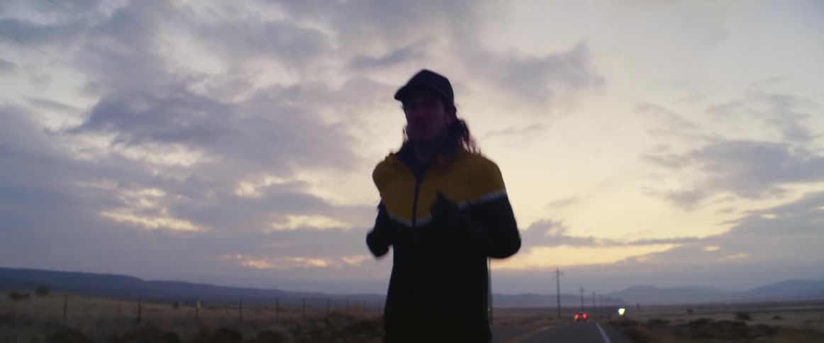 Man with long curly hair and cap running along an asphalt road is silhouetted against the sky