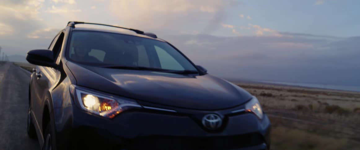 A woman driving a vehicle on a road surrounded by grassy plains with the sky and clouds in the background