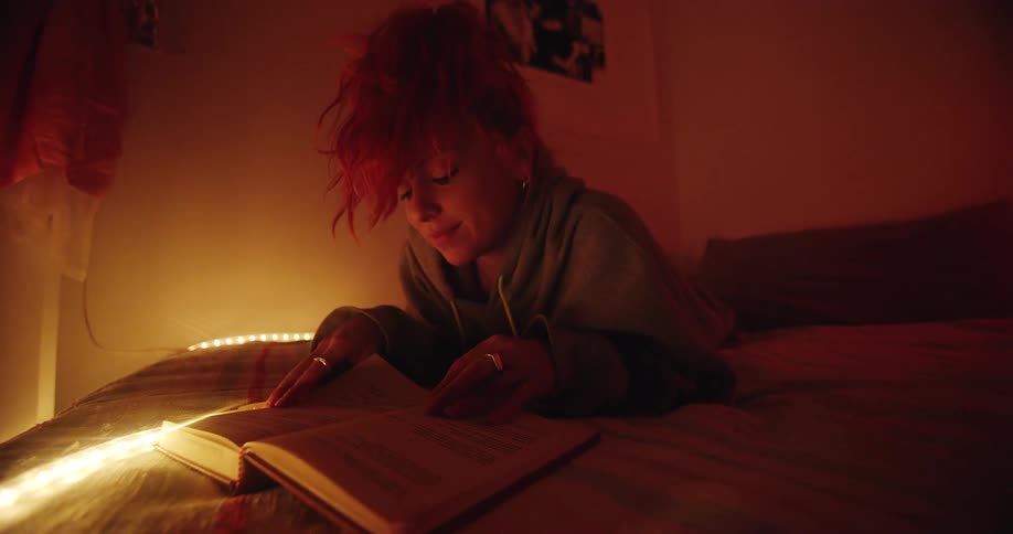 Woman lies on bed in dimly lit room flipping through book
