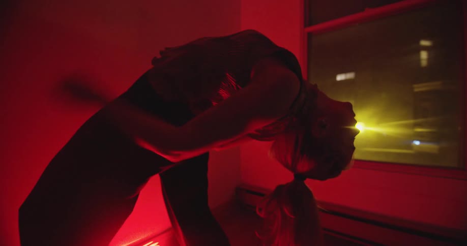 Woman bends her body backwards then returns to a standing position while a red light illuminates the room
