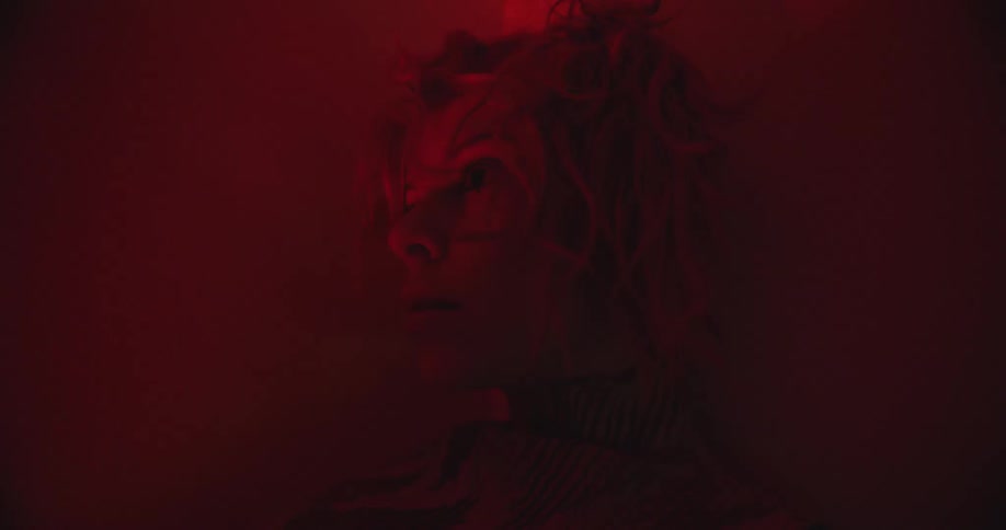 A woman leans strangely up against the wall in a room soaked in red light
