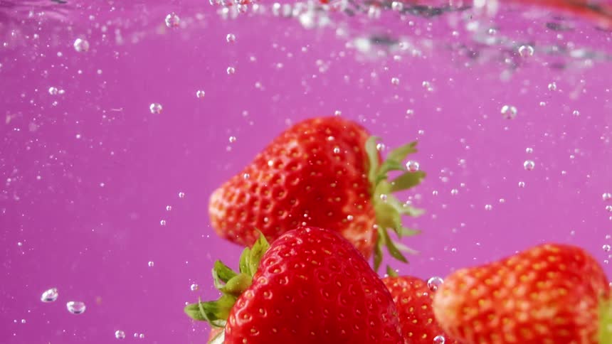 Red ripe strawberries fall into water in front of a pink background