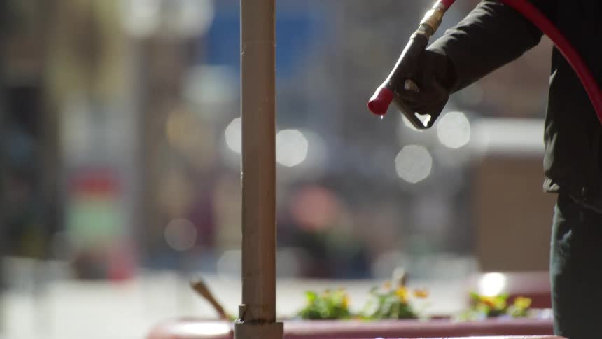 One person points as another wearing gloves waters flowers in large pots near street