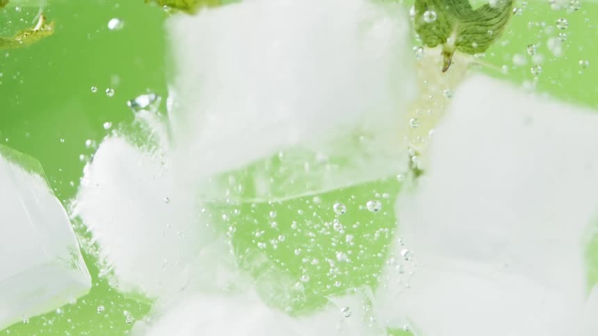 Ice falls into water with lime slices and mint in front of a green background