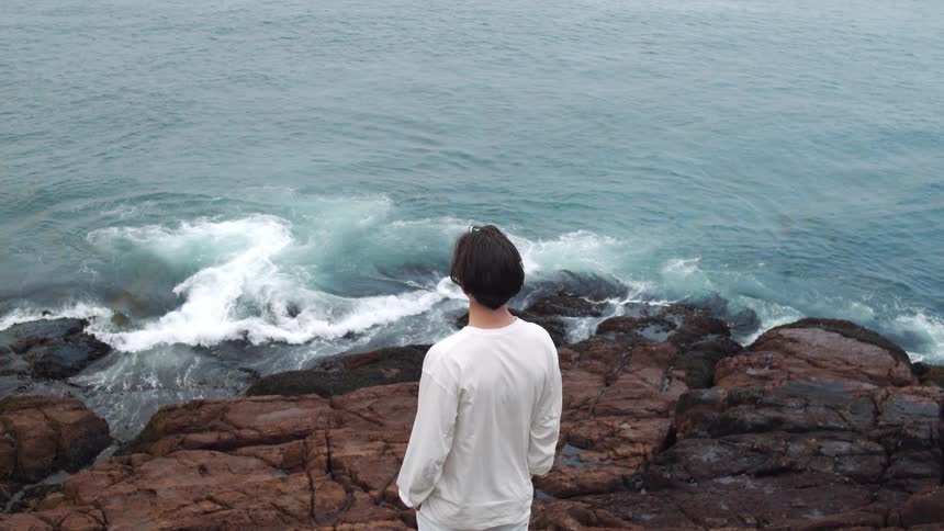Waves wash onto a rock formation on which a man dressed in all white stands looking out over the ocean