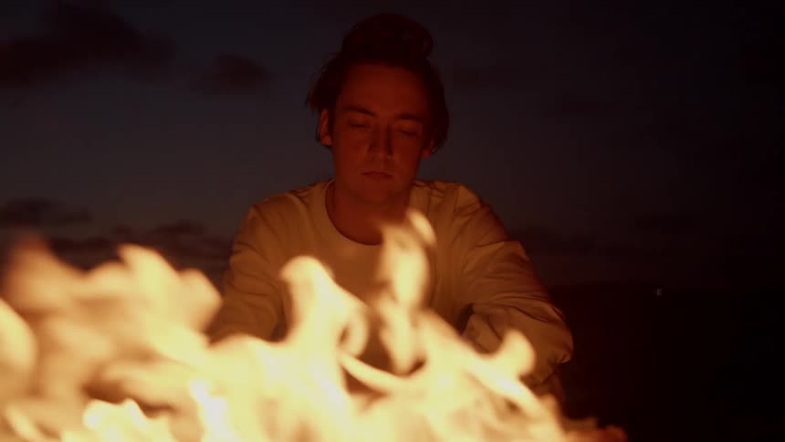 A young man in a white long sleeve t shirt watches a campfire at dusk