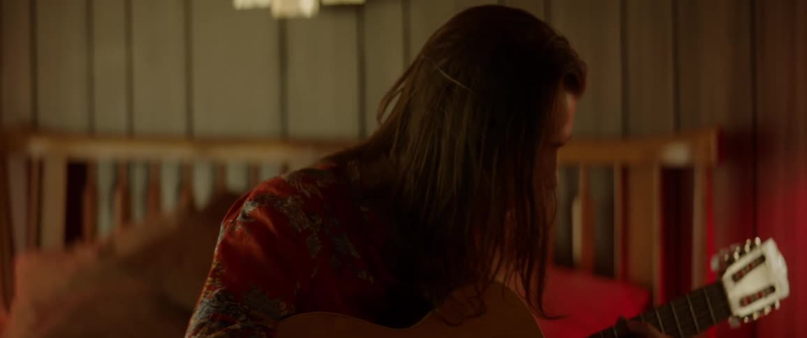 Young man with long hair plays acoustic guitar while sitting on the bed in a motel room