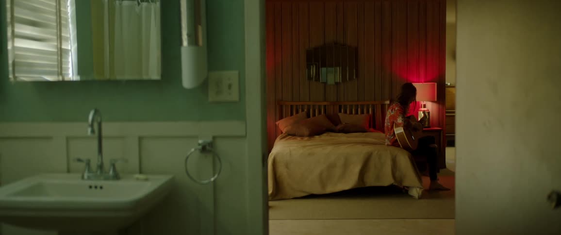 Man with a red shirt sits on a bed and plays a guitar while a woman passes by