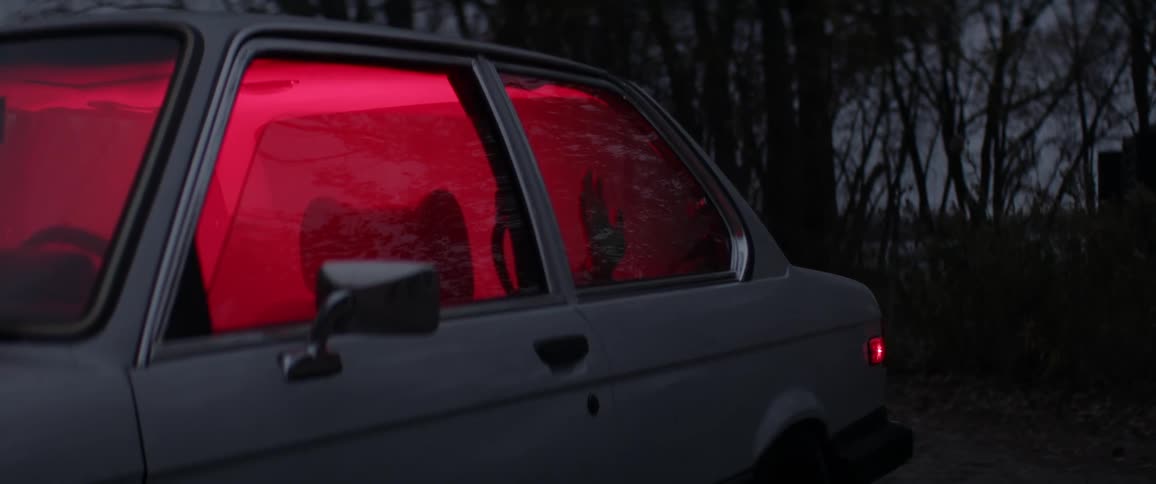 A hand touches the window of a car, filled with smoke and glowing red from inside