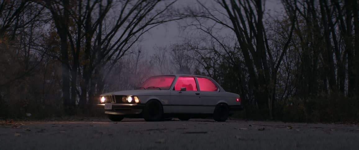 Smoke billows out of a car, glowing red from inside, parked in a country road