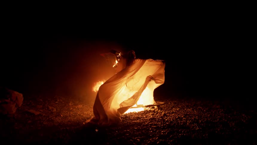 A shopping covered in a white sheet burns on fire on a rocky beach at night