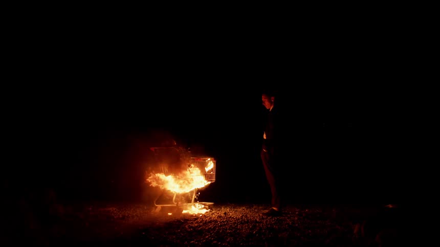 A young man watches a shopping cart burn at night