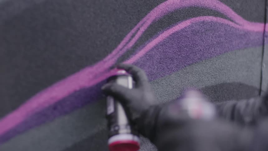 A person wearing a coat and black gloves spray paints a design on a wall