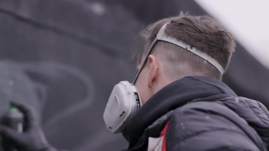 Man wearing a face mask spray paints a graffiti on a wall outside