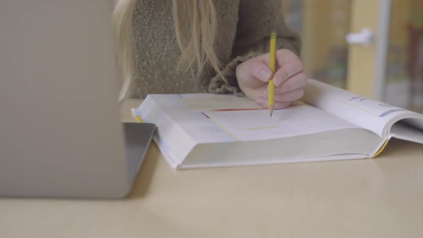 A young woman is reading a text book that lays open next to her laptop as she studies