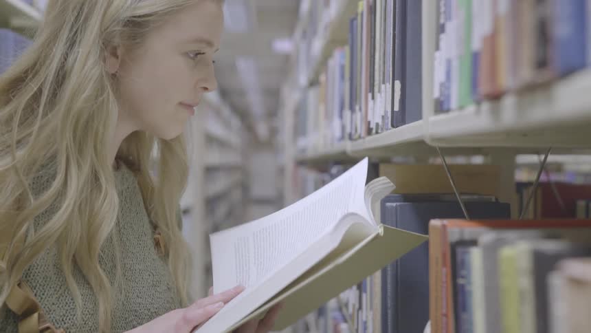 A woman in a library next to bookshelves reading a book with people walking in the background