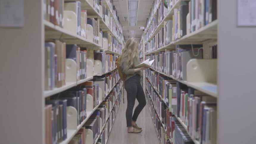 Blonde girl with a grey pullover and a brown backpack looks through a book in the library