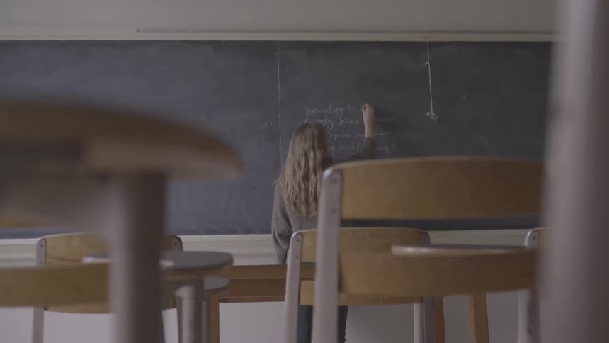 Blonde girl with a grey pullover writes on a blackboard with chalk in the class
