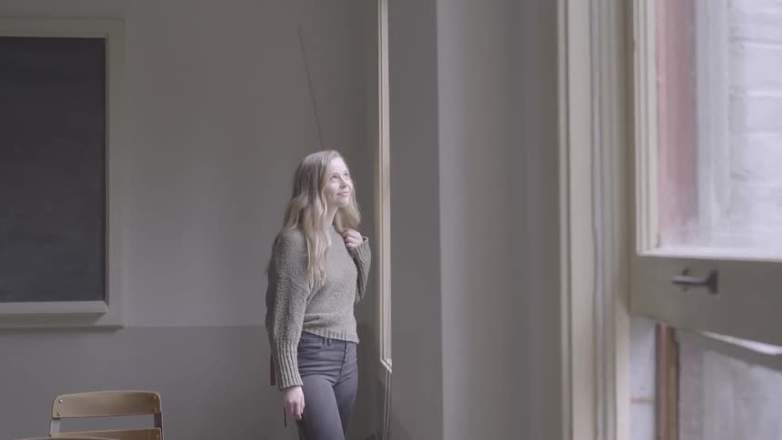A young woman standing near the window in an old fashioned classroom smiles while gazing outside
