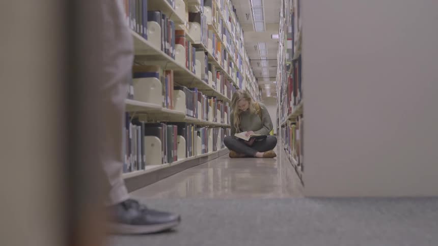 A teenage girl sits in an aisle in a library reading a book