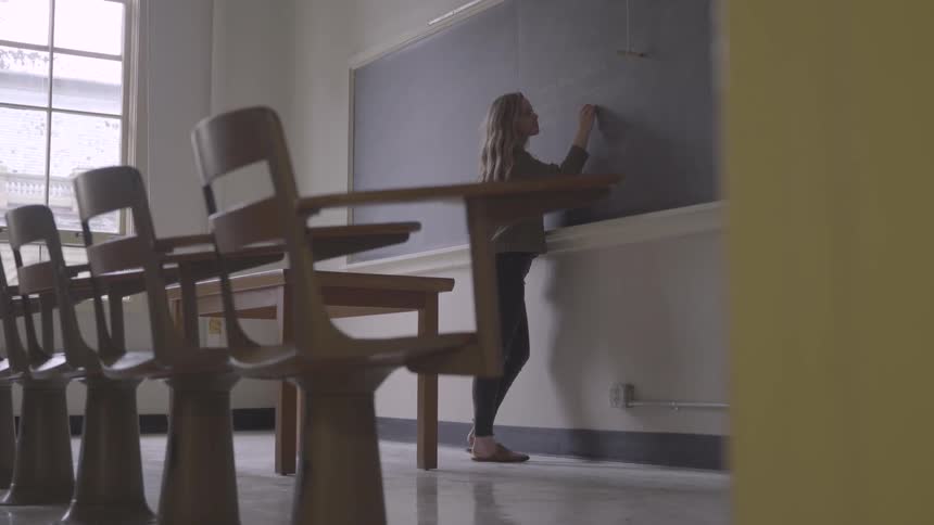 Teacher stands in front of an empty classroom and uses chalk to write on the chalkboard