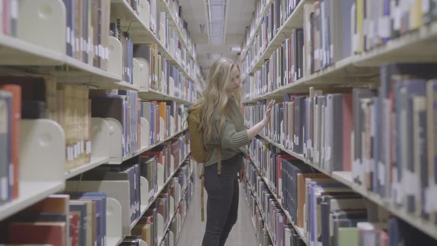 Blonde teenager girl with a grey pullover a brown backpack and pants searches for a book in the public library