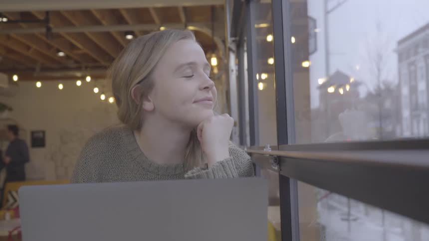 Blonde teenage girl with a pullover smiles slightly and looks out the windows in a cafeteria