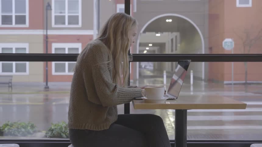 Blonde teenage girl sitting at a table drinks a cup of coffee and uses her laptop in a cafe