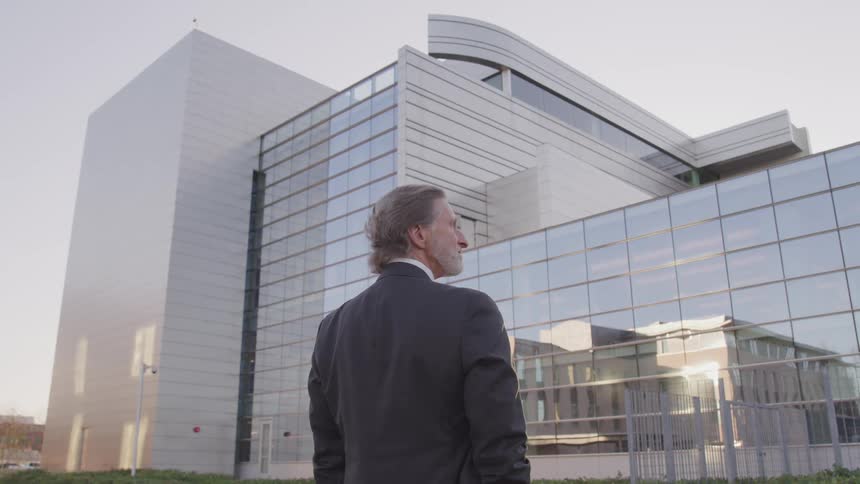 A graying man in a business suit stands with his hands in his pockets outdoors in front of large building with a reflective facade