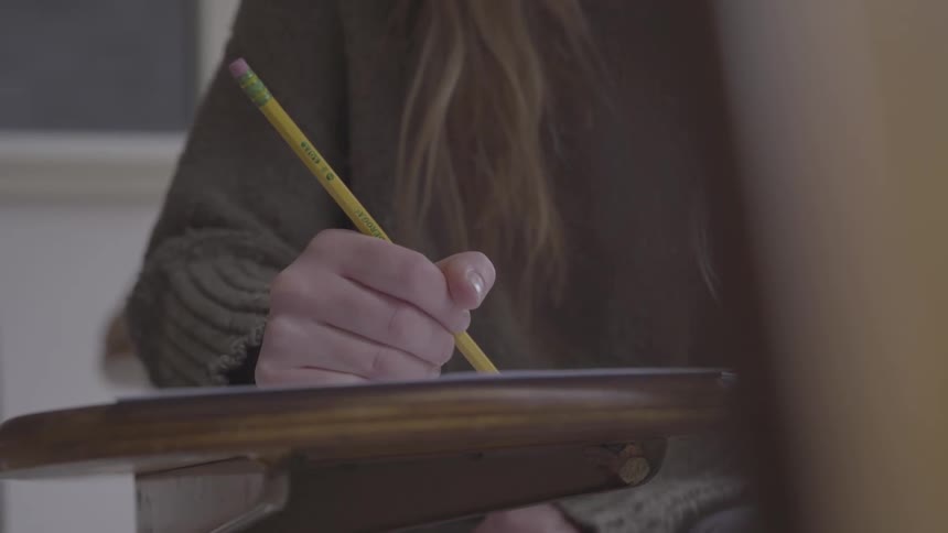 A young woman writes on a sheet of paper with a pencil while sitting at a school desk