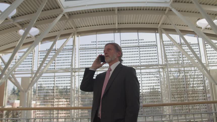 A businessman standing in an atrium is listening to a phone call and then turns to his right
