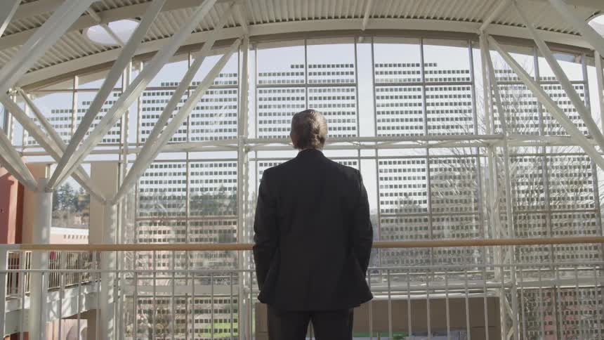 A man with a suit standing next to hand railing with a large window in the background